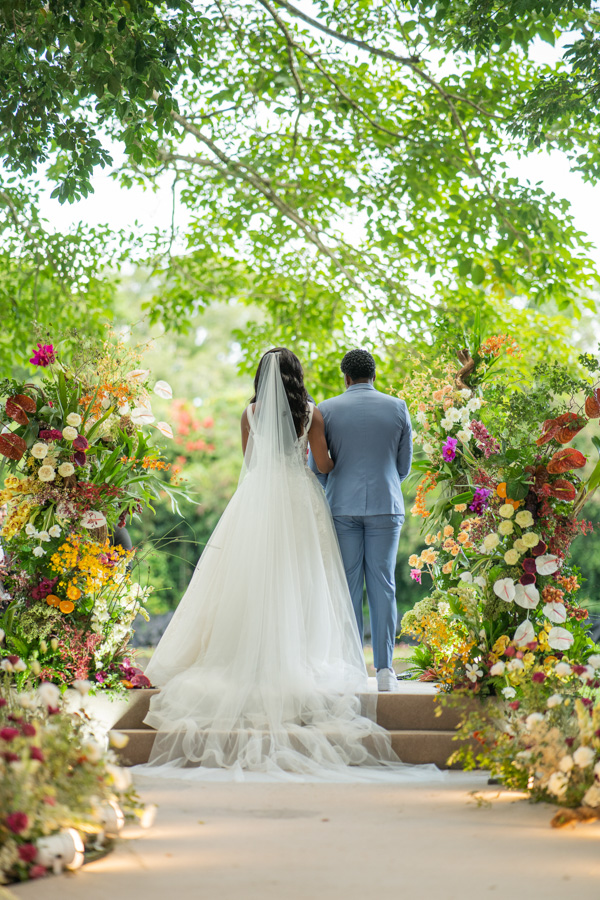 Casamento de Camilla de Lucas e Mateus Ricardo no Rio de Janeiro, foto do casal de costas, na hora da cerimônia, com o fundo de árvores e flores coloridas