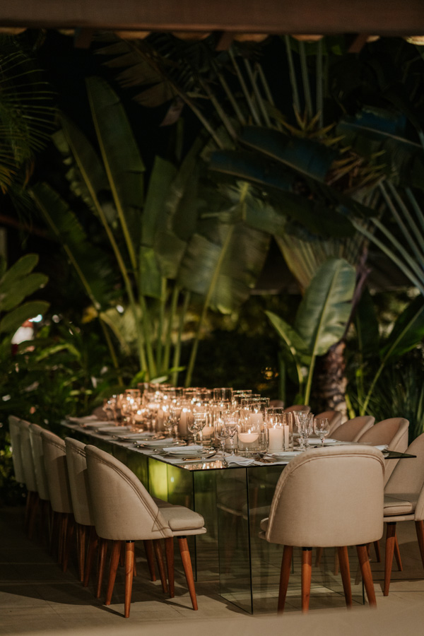 Decoração do noivado de Paula Aziz e Francisco Garcia, cadeiras em marrom claro com o contraste das velas sobrepostas na mesa, fundo verde de folhas, mesa de vidro 