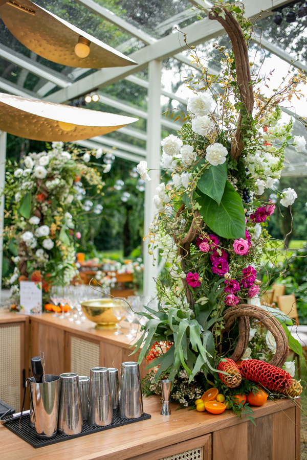 Casamento de Camilla de Lucas e Mateus Ricardo, espaço da mesa de bar para os convidados, taças de bebidas prtas dispostas em cima da mesa, pé de flores rosas, brancas e laranjas em cima da mesa 