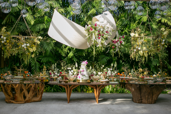 Casamento de Camilla de Lucas e Mateus Ricardo, espaço da mesa do bolo e doce dos noivos, bolo branco com detalhes florais rosa, mesa colorida, fundo com folhas e bolhas de sabão instaladas, espaço de flores penduradas no fundo 