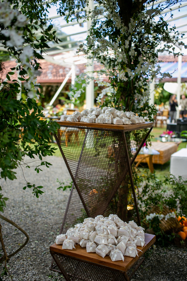 Casamento de Camilla de Lucas e Mateus Ricardo, espaço da lembrancinha para os convidados, lembrancinhas envelopadas por um papel branco com laço destacado, ao fundo flores, folhas e laranjas que enfeitam o local 