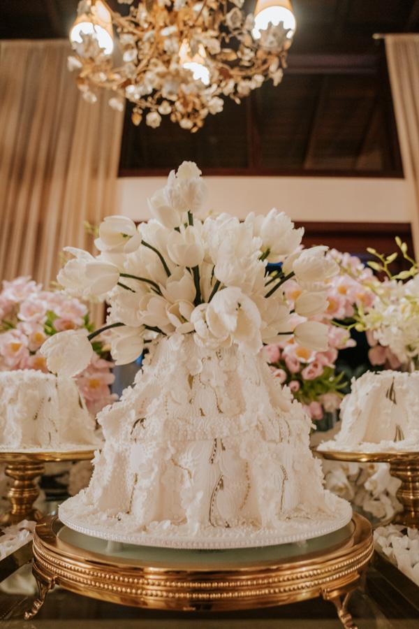 Bolo da mesa de doces do noivado de Paula Aziz e Francisco Garcia, bolo branco com enfeites de tulipa, lustre dourado ao fundo com flores rosas atrás 